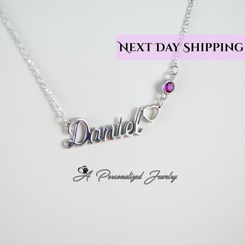 May include: Silver necklace with the name "Daniel" in cursive script, a small heart charm, and a single purple gemstone. The necklace is on a white background with the text "NEXT DAY SHIPPING" and "A Personalized Jewelry" in the bottom corners.