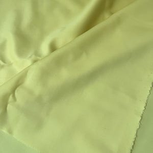 May include: Close-up of a folded piece of light yellow fabric. The fabric has a smooth texture and a slight sheen, with visible folds and creases. The edge of the fabric is unfinished, showing the raw edge of the material. The overall appearance is soft and flowing.