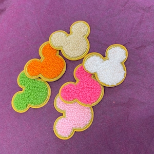 May include: Six colourful felt Mickey Mouse head patches with gold outlines. The patches are in shades of green, orange, pink, and white.