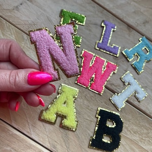 May include: A set of felt letters with gold glitter trim. The letters are in various colours, including pink, green, purple, white, yellow, and black. The letters spell out the word "NEW"