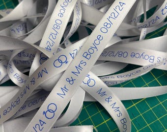 May include: White satin ribbon printed with blue text. The text reads "Mr & Mrs Boyce 08/12/24" along with a graphic of two intertwined rings. The ribbon is scattered across a green surface, ideal for wedding favors or decorations.