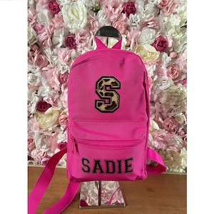 May include: A pink backpack with a leopard print letter "S" and the name "SADIE" embroidered on the front.
