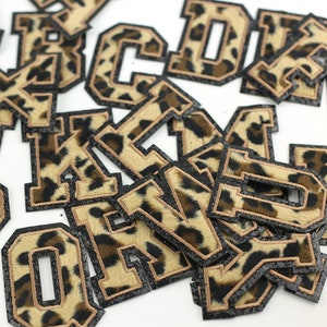 May include: A selection of leopard print letter patches. Each letter features a tan and black spotted pattern with a black border and tan stitching. The letters are in a varsity style.
