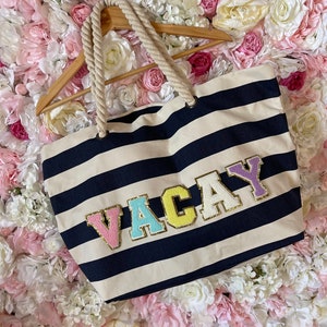May include: A striped navy blue and white canvas tote bag with a rope handle. The bag has the word "VACAY" spelled out in colourful felt letters with gold glitter accents.
