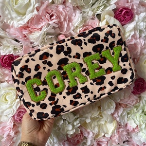 Könnte beinhalten: Eine rosa und braune Kosmetiktasche mit Leopardenmuster und dem Namen "COREY" in Grün gestickt.