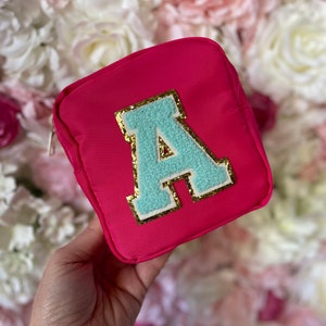 May include: A bright pink zippered pouch with a large turquoise letter A with gold glitter trim. The pouch is held in a hand against a background of pink and white flowers.