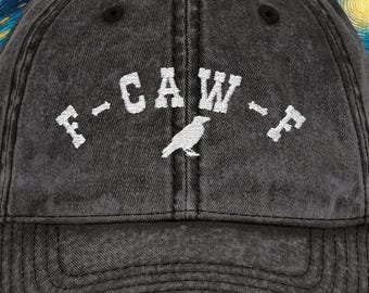 May include: A distressed charcoal gray baseball cap with the text "F-CAW-F" in white, with a small bird graphic. The cap has a curved brim and a classic six-panel design. The background features a starry night sky.