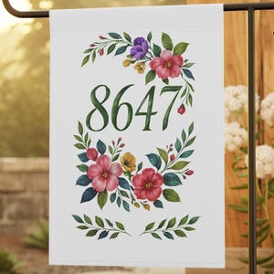 May include: A white garden flag featuring a floral wreath design. The wreath is composed of green leaves and pink, red, and purple flowers. The number 8647 is printed in the centre of the wreath.