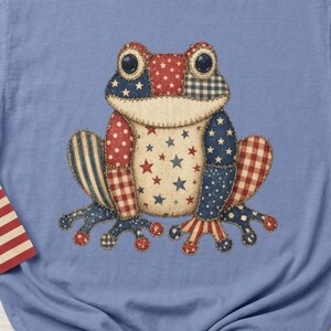 May include: A pale blue t-shirt with a frog design in red, white, and blue patchwork. The frog's design incorporates stars and stripes. An American flag and a star-shaped decoration are also visible in the image.