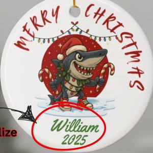 May include: A white ceramic Christmas ornament with a cartoon shark wearing a Santa hat and holding candy canes. The ornament has the words "Merry Christmas" and the name "William" with the year "2025" printed on it. A gold string is attached for hanging.