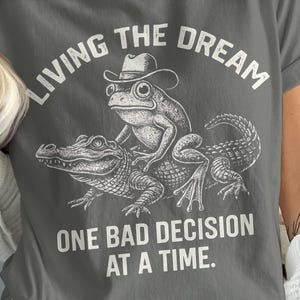 May include: Gray t-shirt with a graphic of a frog wearing a cowboy hat riding an alligator. The text "LIVING THE DREAM" arches above the image, with "ONE BAD DECISION AT A TIME" below. The shirt is a casual, unisex style.