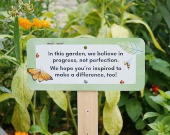 May include: A green sign with a white border and rounded corners is staked in the ground. The sign has a butterfly, a bee, and text that reads: "In this garden, we believe in progress, not perfection. We hope you're inspired to make a difference, too!"