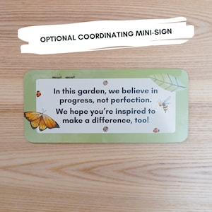 K&ouml;nnte beinhalten: Ein rechteckiges gr&uuml;nes Mini-Schild mit abgerundeten Ecken, das den Text "In this garden, we believe in progress, not perfection. We hope you're inspired to make a difference, too!" enth&auml;lt. Dekorative Elemente sind ein Schmetterling, Marienk&auml;fer, Ameisen, eine Biene und ein Blatt.