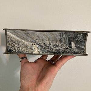 May include: A hand-painted book cover with a black and white landscape scene. The scene depicts a path leading to a house with a fence and trees in the background.