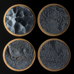 May include: Four round coasters with a wood rim and a 3D printed design of a city map. The coasters are black and grey.