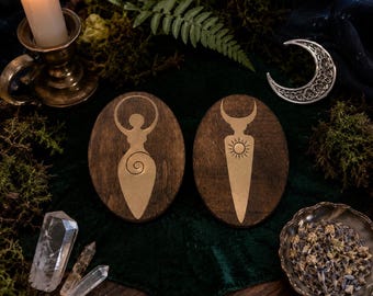 Puede incluir: Dos placas ovaladas de madera con grabados de color dorado. La placa izquierda muestra una figura femenina estilizada con una espiral en el vientre, mientras que la derecha presenta una luna creciente sobre un símbolo solar. Sobre tela verde.