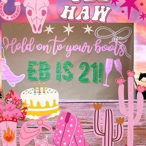 May include: A pink and green sign with the text "Hold on to your boots EB is 21!" with a pink cowboy hat, a horseshoe, and a lasso. There is a pink cactus, a pink cowboy boot, and a pink bandana on a wooden surface.