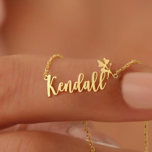 May include: Gold necklace with the name "Kendall" spelled out in cursive script. A small fairy charm is attached to the end of the name.