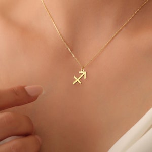 May include: A delicate gold-coloured necklace featuring a Sagittarius zodiac symbol pendant. The pendant is a stylized arrow and cross design. The necklace is worn against a white top.