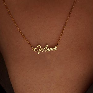 May include: A delicate gold necklace with the word "Mama" in cursive script. The necklace is displayed on a person's neck, with the chain and pendant clearly visible. The pendant is a stylish and sentimental piece of jewelry.
