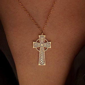 May include: A gold-coloured Celtic cross pendant necklace. The pendant features an intricate knot design within the cross shape. The necklace chain is delicate and also gold-coloured. The necklace is displayed against a dark background.