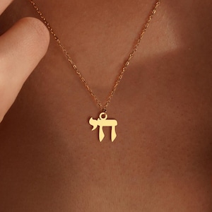 Peut inclure: Un collier dor&eacute; avec une cha&icirc;ne d&eacute;licate et un pendentif en forme de la lettre h&eacute;bra&iuml;que "Chai". Le pendentif est centr&eacute; sur la poitrine. Le collier est port&eacute; sur un fond de couleur chair.