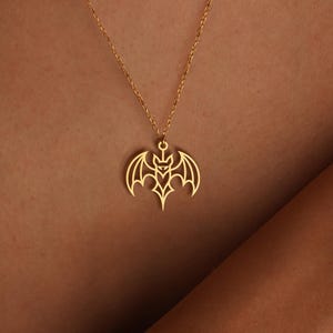 K&ouml;nnte beinhalten: Eine goldfarbene Halskette mit einem Fledermaus-Anh&auml;nger. Der Fledermaus-Anh&auml;nger hat ein detailliertes, offenes Design mit ausgebreiteten Fl&uuml;geln. Die Halskette wird &uuml;ber einem gr&uuml;nen Oberteil getragen.