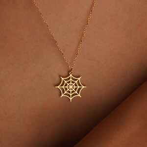 May include: A gold-colored necklace featuring a spiderweb pendant. The pendant is a detailed, openwork design, suspended from a delicate chain. The necklace is worn against a green top.