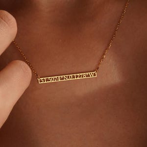 May include: A gold-colored necklace with a delicate chain and a horizontal bar pendant. The bar features the engraved coordinates "51.5074° N 0.1278° W". The necklace is worn around the neck.