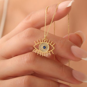 May include: Gold necklace featuring a blue evil eye pendant. The pendant is a detailed outline of an eye with a blue gemstone at its centre.