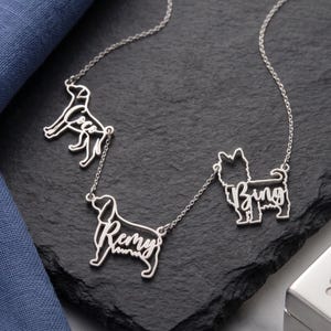 May include: A silver-toned necklace featuring three distinct dog-shaped pendants, each integrated with a name in cursive script. The pendants are labeled with the names Coco, Remy, and Bing, connected by a delicate silver chain link design.