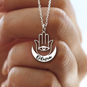 May include: Silver necklace with a hamsa hand charm and a crescent moon with the name "Olivia" inscribed on it.