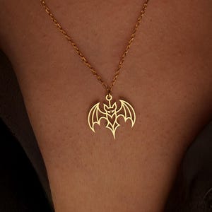 K&ouml;nnte beinhalten: Eine goldfarbene Halskette mit einem Fledermaus-Anh&auml;nger. Der Fledermaus-Anh&auml;nger hat ein detailliertes Design mit ausgebreiteten Fl&uuml;geln und einem stilisierten Gesicht. Die Halskette wird vor einem dunklen Hintergrund getragen.
