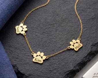 May include: A gold-toned necklace featuring three paw print charms connected by a delicate chain. Each charm is engraved with a name: Neal, Kim, and Phill. The metal charms have a polished finish and are spaced evenly along the length of the chain.