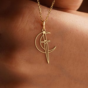 May include: A gold-colored necklace featuring a crescent moon and sword pendant. The pendant is a delicate, open-work design. The chain is a fine link style. The necklace is displayed against a blurred background.