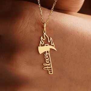 K&ouml;nnte beinhalten: Eine goldfarbene Halskette mit Anh&auml;nger, die ein Feuerwehrbeil-Design mit Flammen und dem Namen "Alexis" in Schreibschrift zeigt. Die Halskette h&auml;ngt an einer zarten Kette.