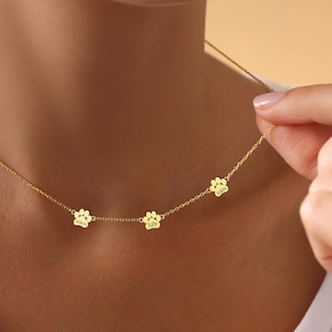 May include: A gold necklace with three paw print charms, each engraved with a name: "Lola", "Lily", and "Sadie".