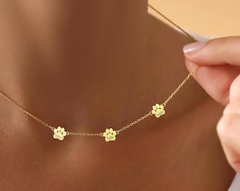May include: A gold-toned chain necklace featuring three small, evenly spaced paw print charms. Each charm is engraved with a name, specifically Willow, Lily, and Eddie, suspended on a delicate link chain that rests against the collarbone area.