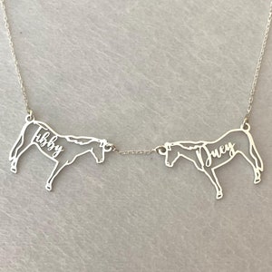 May include: A silver necklace featuring two horse pendants connected by a delicate chain. Each horse silhouette has a name inscribed within: "Libby" and "Lucy". The necklace is displayed on a light gray surface.