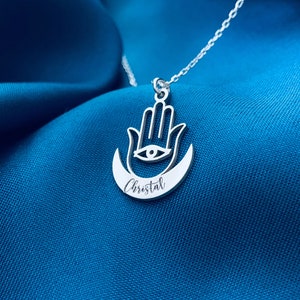 May include: Silver necklace with a hamsa hand charm and a crescent moon. The charm has an eye in the center and the name "Cristal" is engraved on the moon.