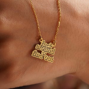 May include: A gold-colored necklace with a pendant that has the words 'fuck around & find out' in a stacked, bold font. The necklace chain is delicate, and the pendant is attached to the chain with small loops. The necklace is displayed against a skin-toned background.
