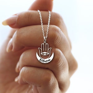 May include: Silver necklace with a hamsa hand charm and a crescent moon with the name Olivia inscribed on it.