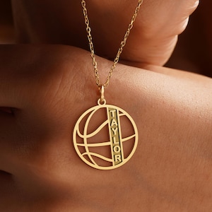 Puede incluir: Un collar dorado con un colgante con forma de bal&oacute;n de baloncesto. El colgante tiene un dise&ntilde;o de baloncesto con el nombre "TAYLOR" inscrito verticalmente. El collar se muestra sobre un fondo borroso.