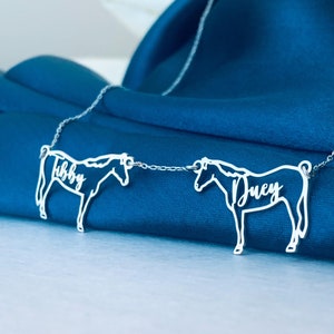 May include: A silver necklace with two horse-shaped pendants linked by a fine chain. The pendants are inscribed with the names "Libby" and "Duey". The necklace is presented on a dark blue fabric.