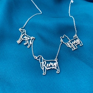 Puede incluir: Un collar de plata con tres siluetas de perros, cada una con un nombre inscrito en su interior. Los nombres son "Coco", "Remy" y "Bingo".
