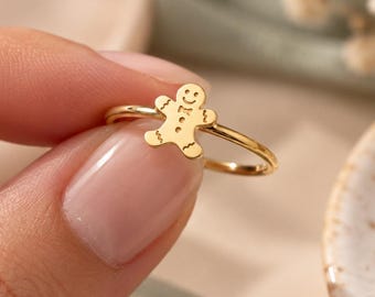 May include: A gold-toned ring featuring a small, flat gingerbread man charm. The charm has engraved facial features, including eyes, a smile, and a bow tie detail. The band is thin and circular, polished to a smooth, reflective finish throughout.