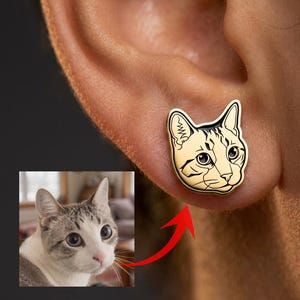 May include: A gold-toned stud earring shaped like a cat head with black engraved line details. The earring features a stylised illustration of a cat face with pointed ears, whiskers, and large eyes, matching the cat shown in the small inset photo.