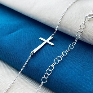 Op de afbeelding: Een zilveren ketting met een delicate zilveren kruis hanger. De ketting wordt getoond op een witte en blauwe stoffen achtergrond.