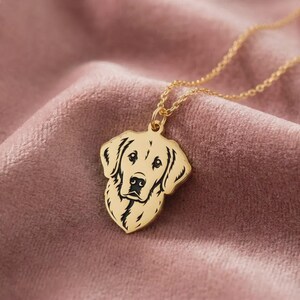 May include: A gold-colored pendant necklace featuring a detailed silhouette of a dog's head. The dog's face is black, and the pendant hangs from a delicate gold chain. The necklace is displayed on a soft, pink velvet surface.