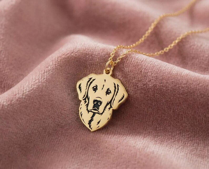 May include: A gold-colored pendant necklace featuring a detailed silhouette of a dog's head. The dog's face is black, and the pendant hangs from a delicate gold chain. The necklace is displayed on a soft, pink velvet surface.